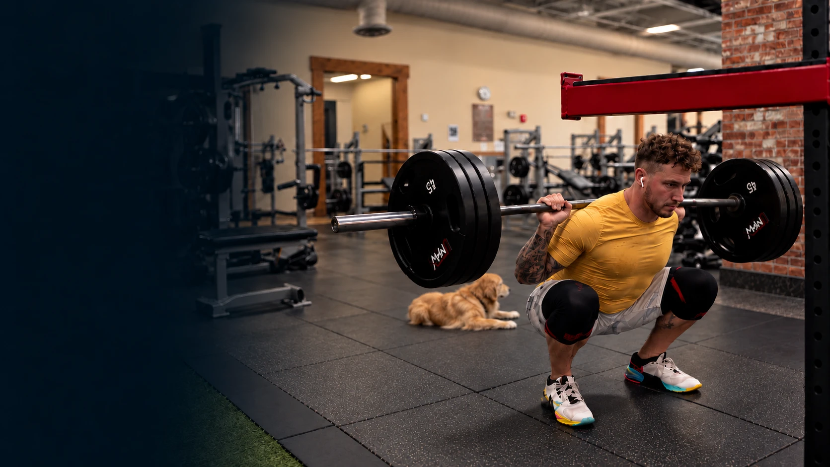 Jeremy Bates squatting with a barbell in the gym, with Zoey resting nearby